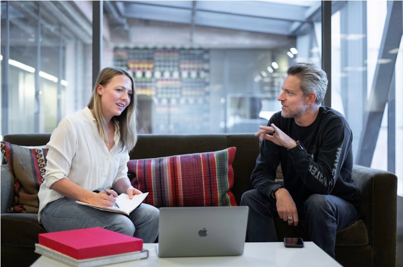A man and woman sit on a couch with a laptop, discussing knowledge management for innovation and continuous improvement.